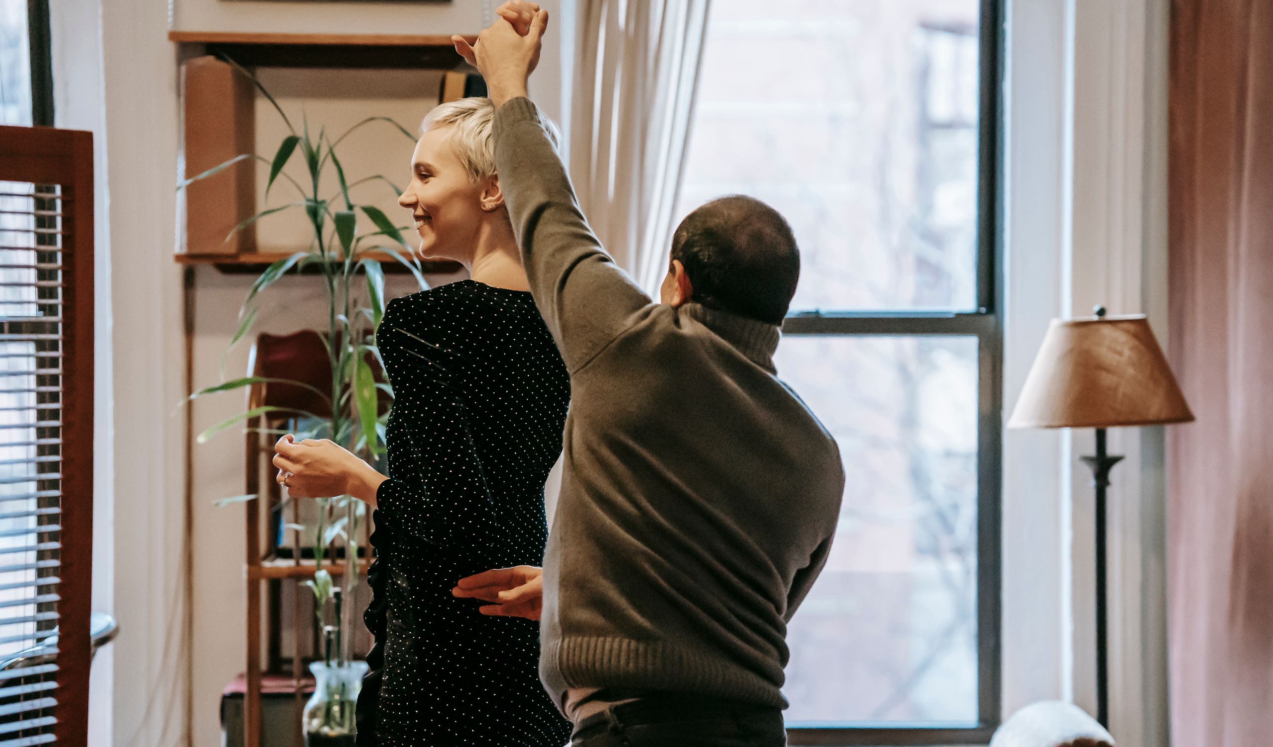Back view of happy couple dancing and holding hands in bright flat near windows and curtains near lamp and plant with decorative elements