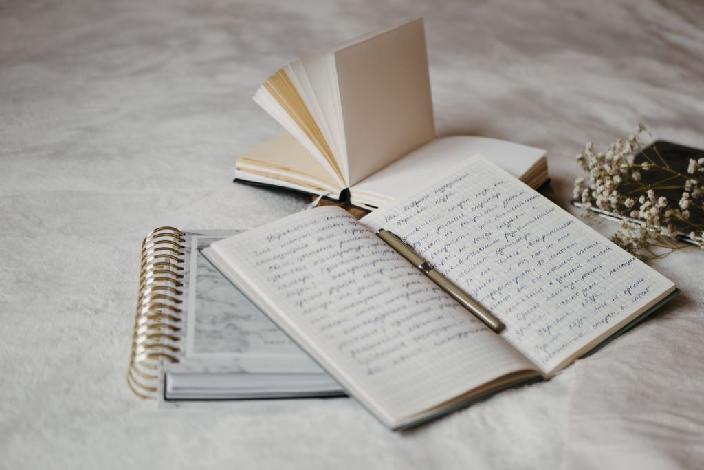 Close-up of open notebooks and a pen on a soft bed surface with delicate dried flowers.