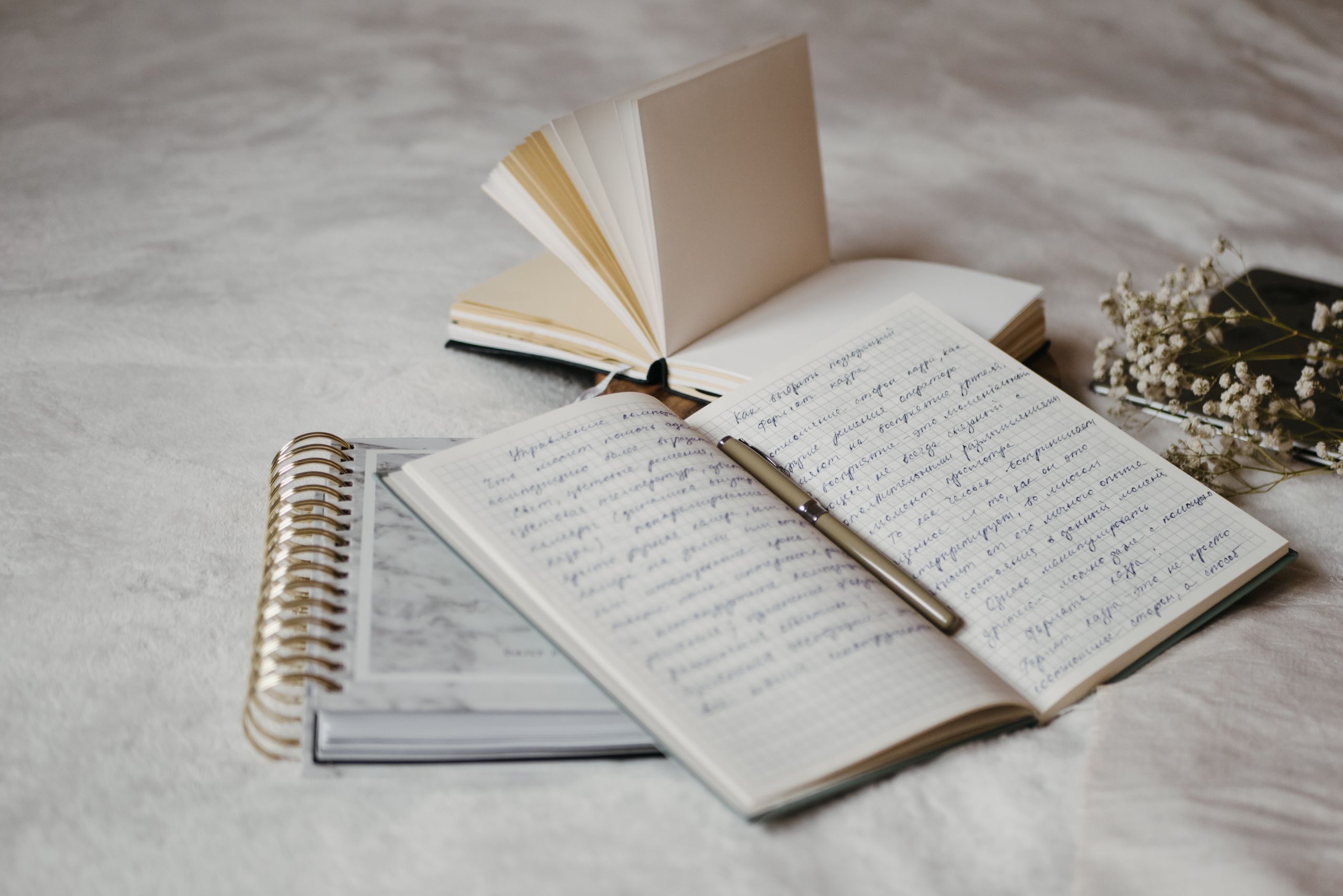Close-up of open notebooks and a pen on a soft bed surface with delicate dried flowers.